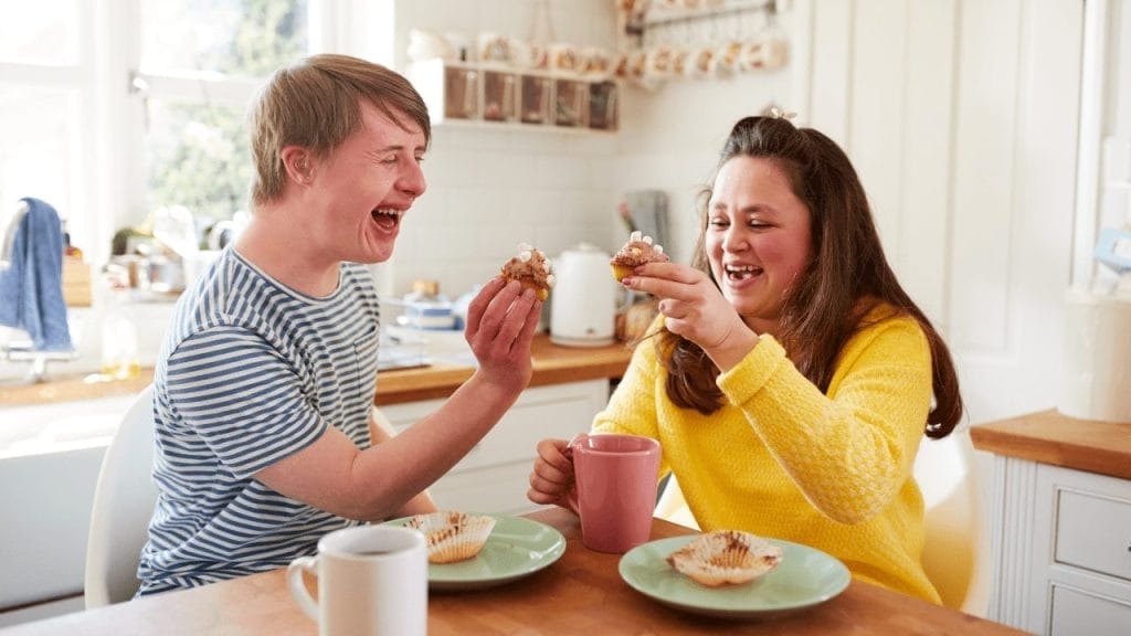 A boy with Down Syndrome eats and laughs with a friend in the kitchen.